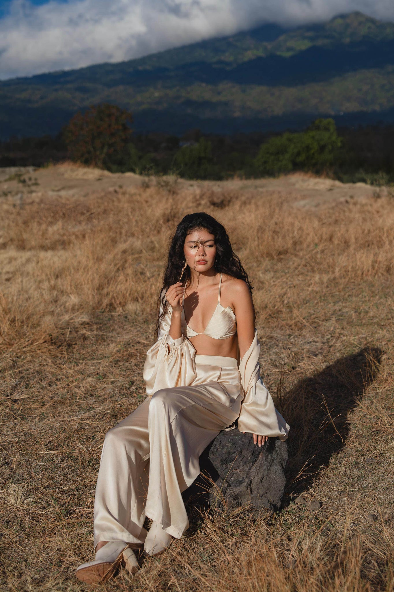 A woman with long, wavy hair and a thoughtful expression sits on a rock in a dry grassy field, wearing the Butterfly Silk Bra in cream by AYA Sacred Wear, with mountains and clouds visible in the background.