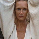 A person with wavy blonde hair and sandy skin wears the Tatva Dress Ivory Colour – Sustainable Bohemian Dress by AYA Sacred Wear, featuring voluminous sleeves, outdoors with arms raised behind their head, gazing intensely at the camera amid greenery.