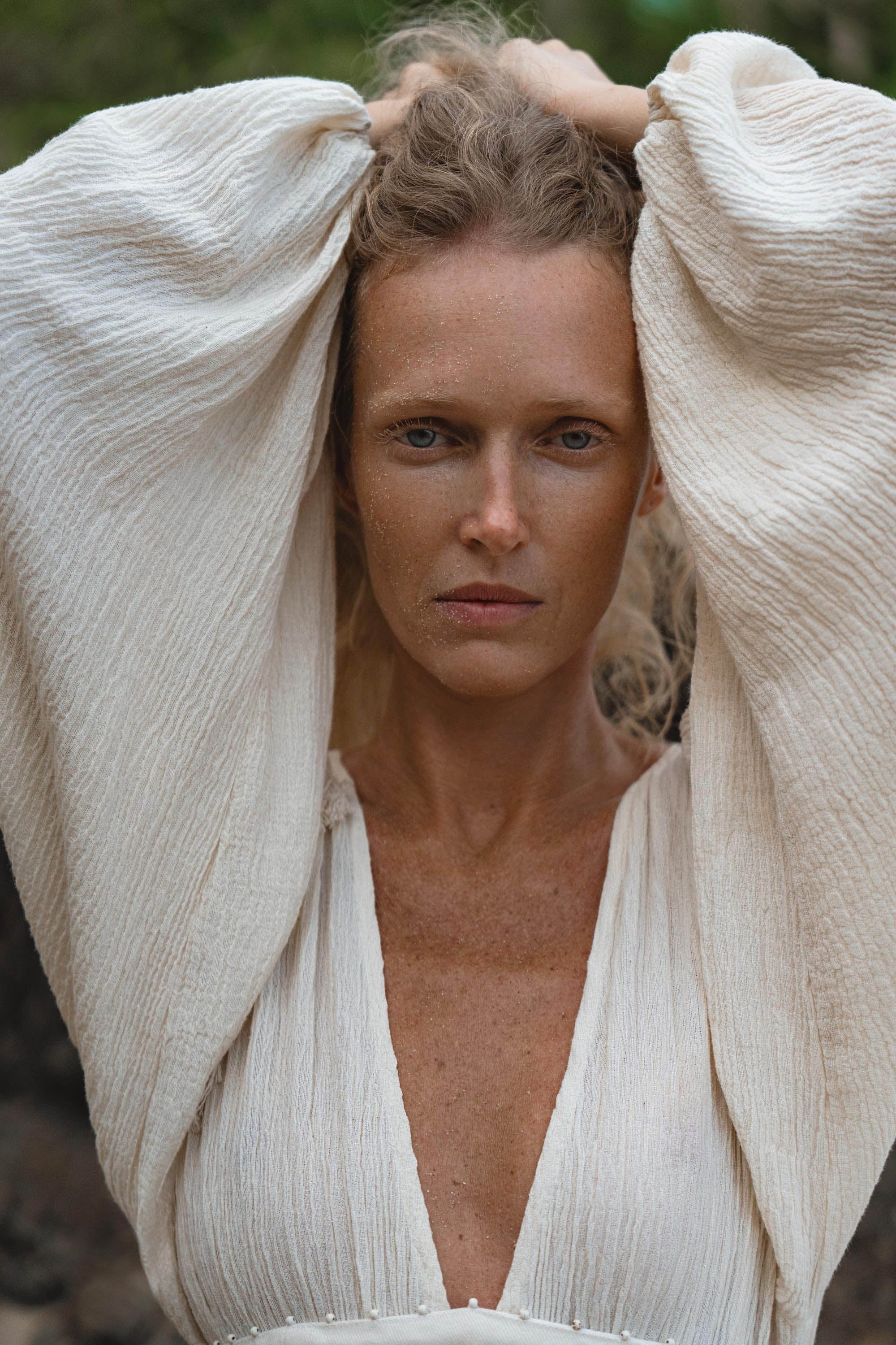 A person with wavy blonde hair and sandy skin wears the Tatva Dress Ivory Colour – Sustainable Bohemian Dress by AYA Sacred Wear, featuring voluminous sleeves, outdoors with arms raised behind their head, gazing intensely at the camera amid greenery.