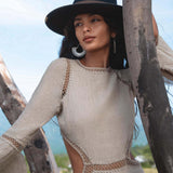 A woman wearing the AYA Sacred Wear Cotton Maxi Dress in Light Sage with long sleeves and a flowing bohemian silhouette stands outdoors near tree branches, gazing thoughtfully into the distance.