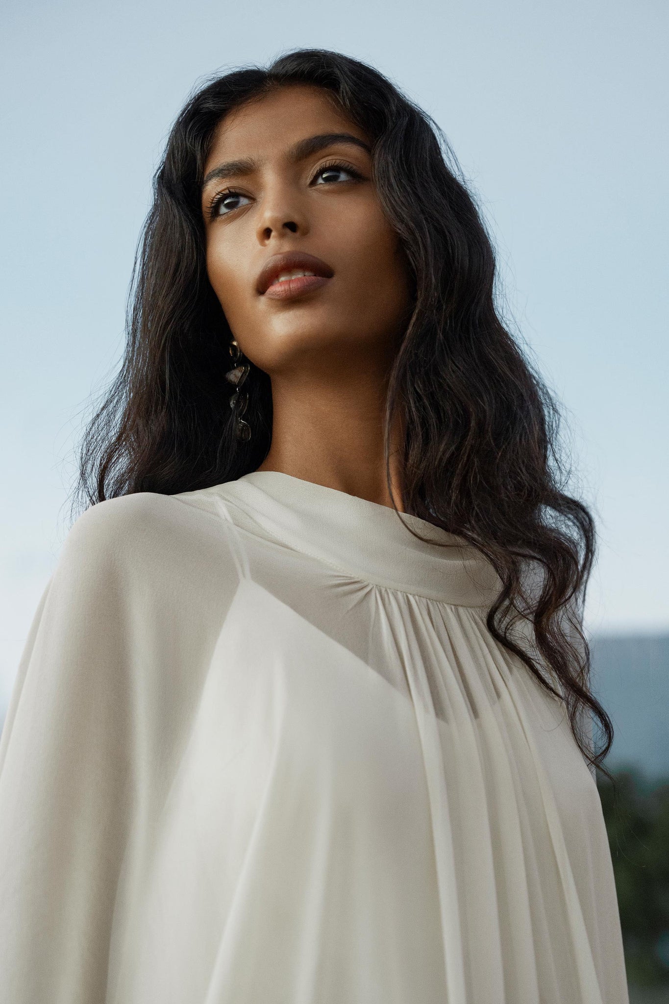 A woman with long wavy dark hair wears the Mahadevi Dress Cream Colour, a pure silk chiffon goddess gown by AYA Sacred Wear, gazing confidently into the distance against a soft blue sky backdrop.