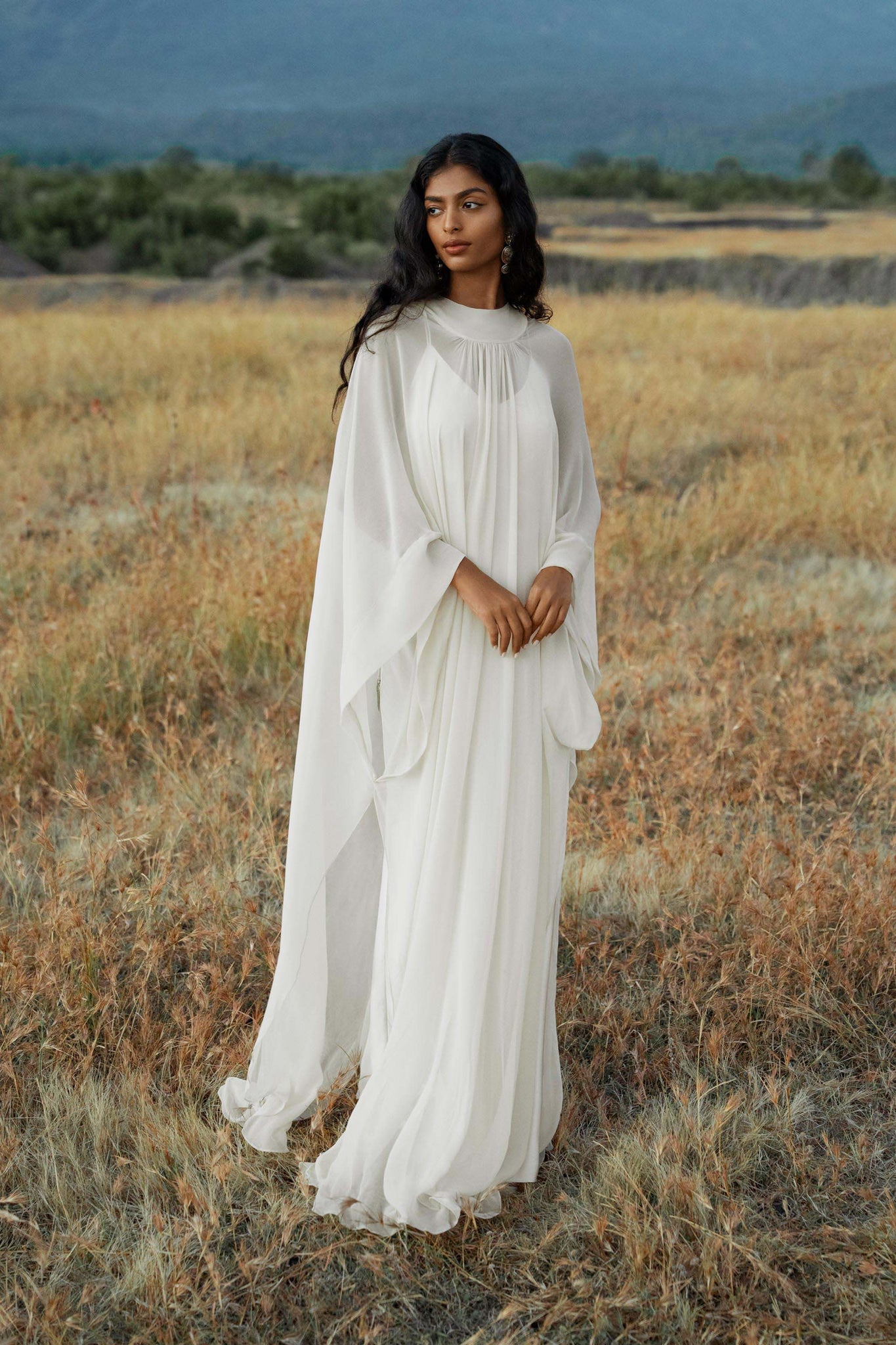 A woman in the AYA Sacred Wear Mahadevi Dress Cream Colour – Pure Silk Chiffon Goddess Gown stands in a dry grassy field with distant mountains, gazing calmly to the side.