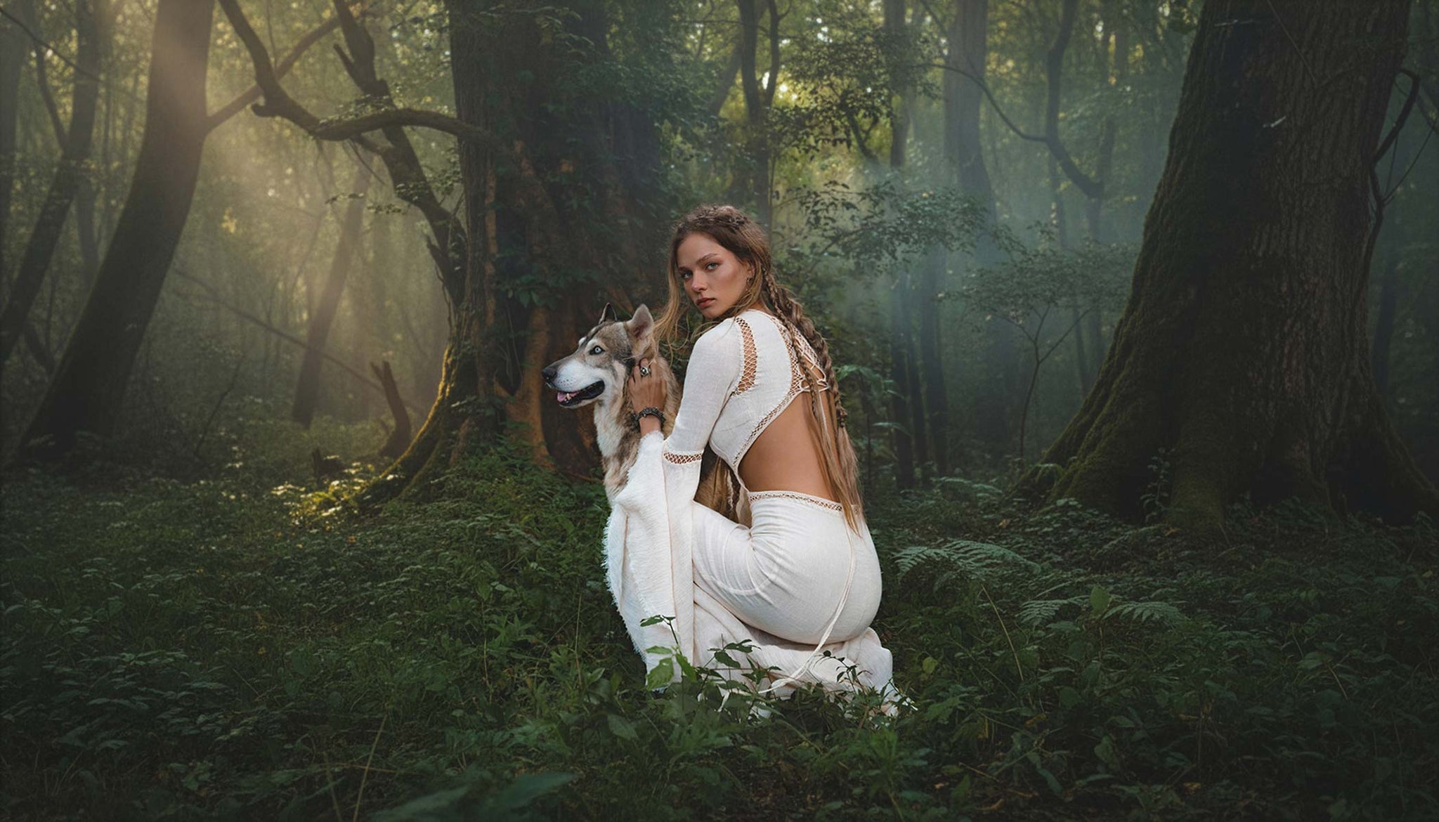 Woman in a white dress kneeling in a forest with a wolf by her side wearing AYA Sacred Wear New 2026 ĀNANDA SHAKTI SS 2026 Organic Collection. 