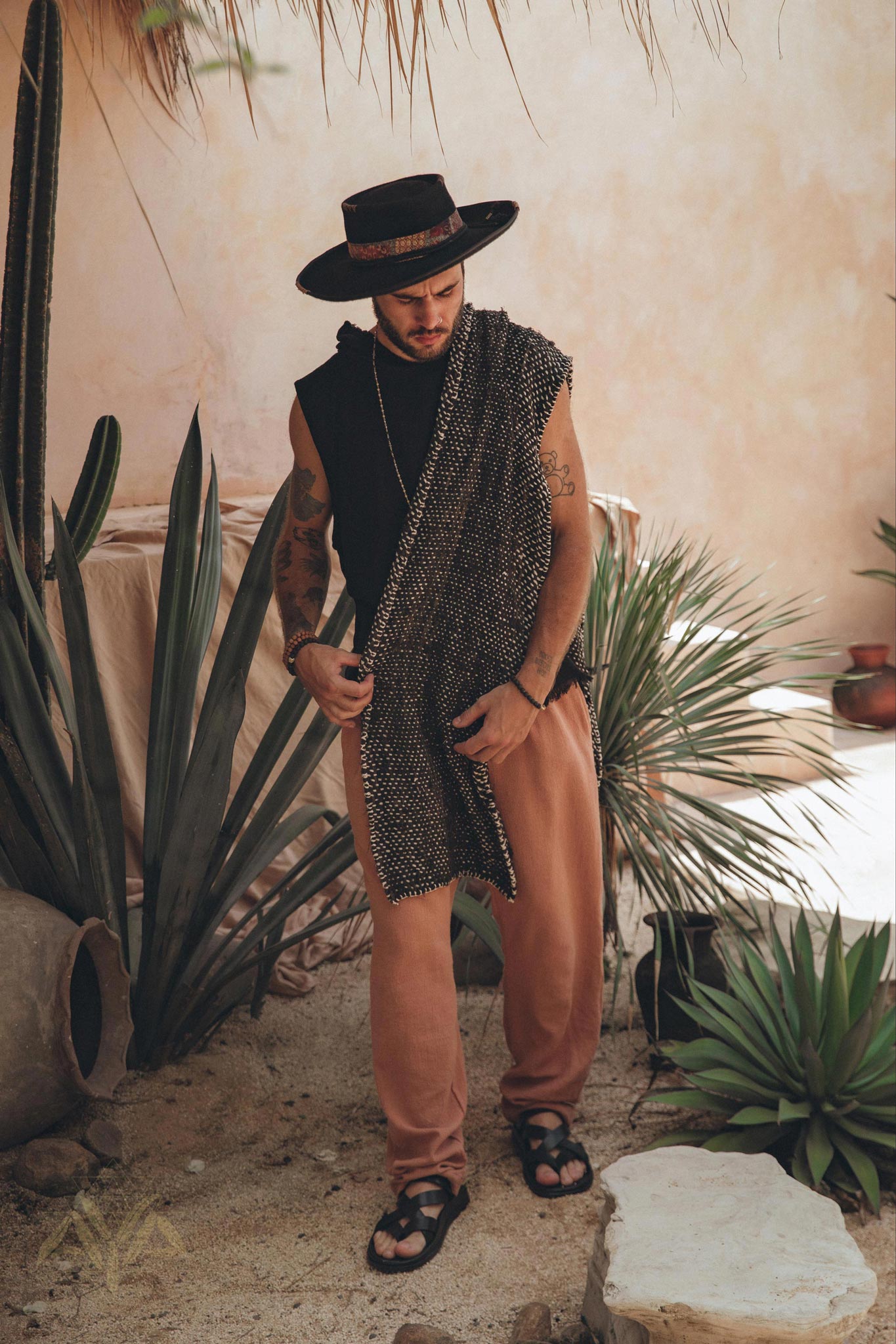 In a rustic, tropical setting brimming with desert plants along a textured stone path, an individual exudes a relaxed and fashionable vibe. They sport a hat, sleeveless shirt, and loose pants, elegantly accessorized with the Black with White Hand-knitted Cotton Scarf for Men by AYA Sacred Wear, dyed botanically.