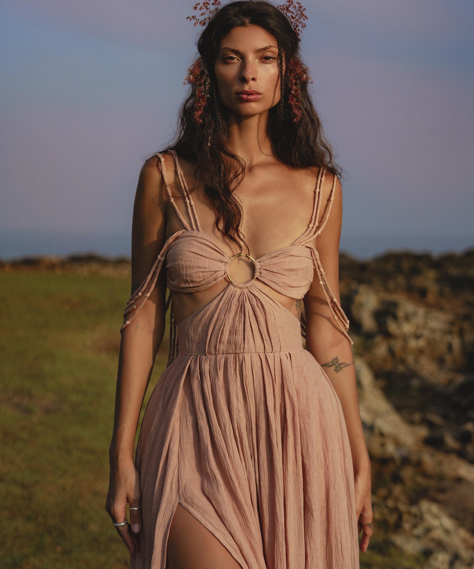 A woman stands on grassy terrain near rocks, wearing the Ananda Lālita Dress Blush Rose – Crinkle Linen Goddess Dress by AYA Sacred Wear. Her long brown hair shines in the sun, adorned with flowers and handcrafted brass rings under a pastel sky.