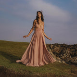 A woman in the Ananda Lālita Dress Blush Rose – Crinkle Linen Goddess Dress by AYA Sacred Wear stands on grassy terrain near rocks, wearing handcrafted brass rings and a floral headpiece, gazing upward with an ethereal, serene presence.