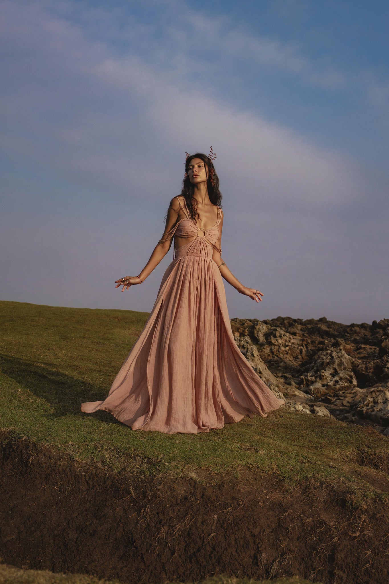 A woman in the Ananda Lālita Dress Blush Rose – Crinkle Linen Goddess Dress by AYA Sacred Wear stands on grassy terrain near rocks, wearing handcrafted brass rings and a floral headpiece, gazing upward with an ethereal, serene presence.