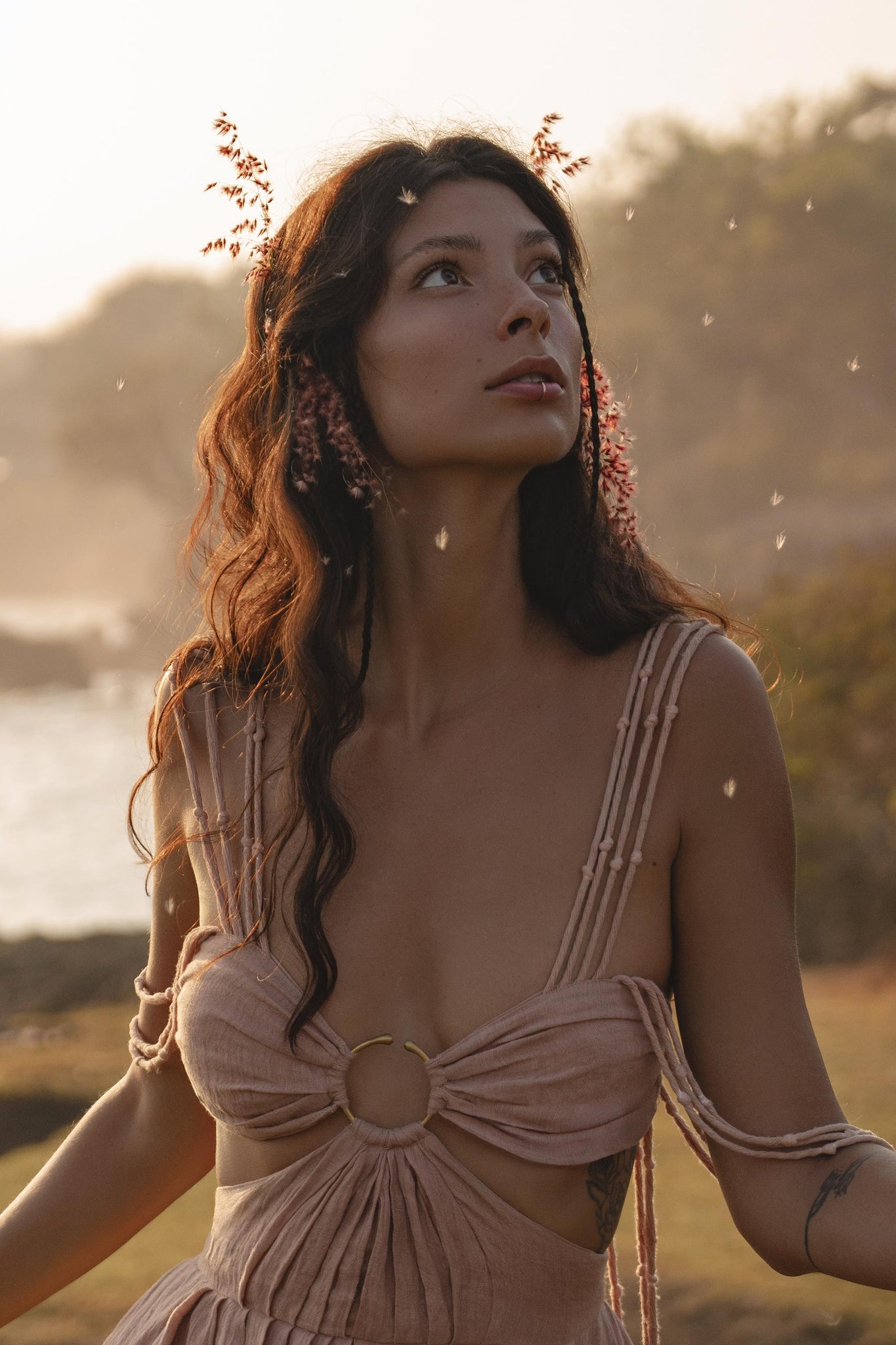 A woman with long wavy brown hair and small pink flowers gazes upward, wearing the Ananda Lālita Dress Blush Rose – Crinkle Linen Goddess Dress by AYA Sacred Wear. Soft sunlight and floating seeds create a dreamy outdoor scene.