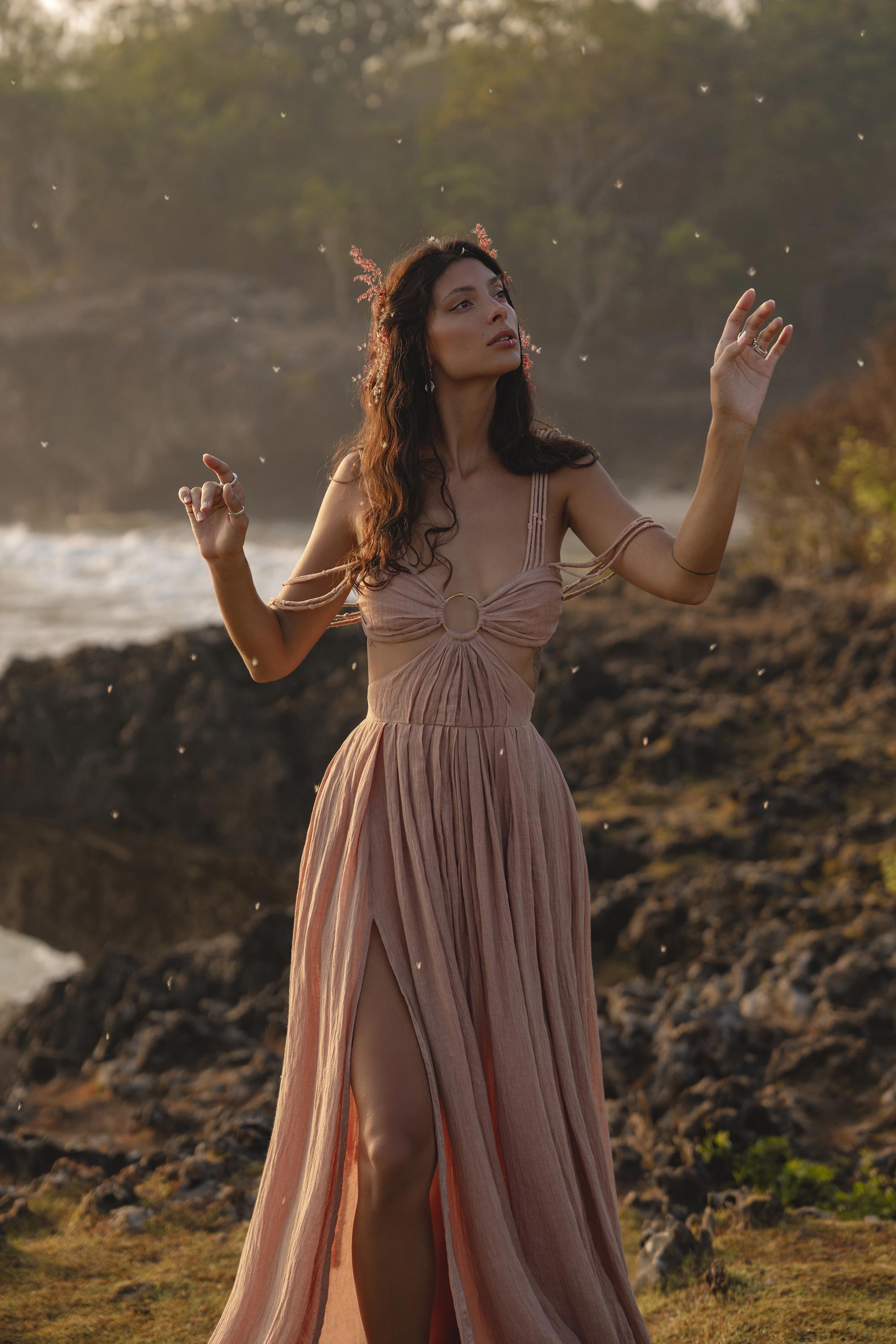 A woman wears the Ananda Lālita Dress Blush Rose – Crinkle Linen Goddess Dress by AYA Sacred Wear, standing on rocky terrain near water, arms raised beneath falling particles and soft sunlight, exuding an ethereal, mystical aura.
