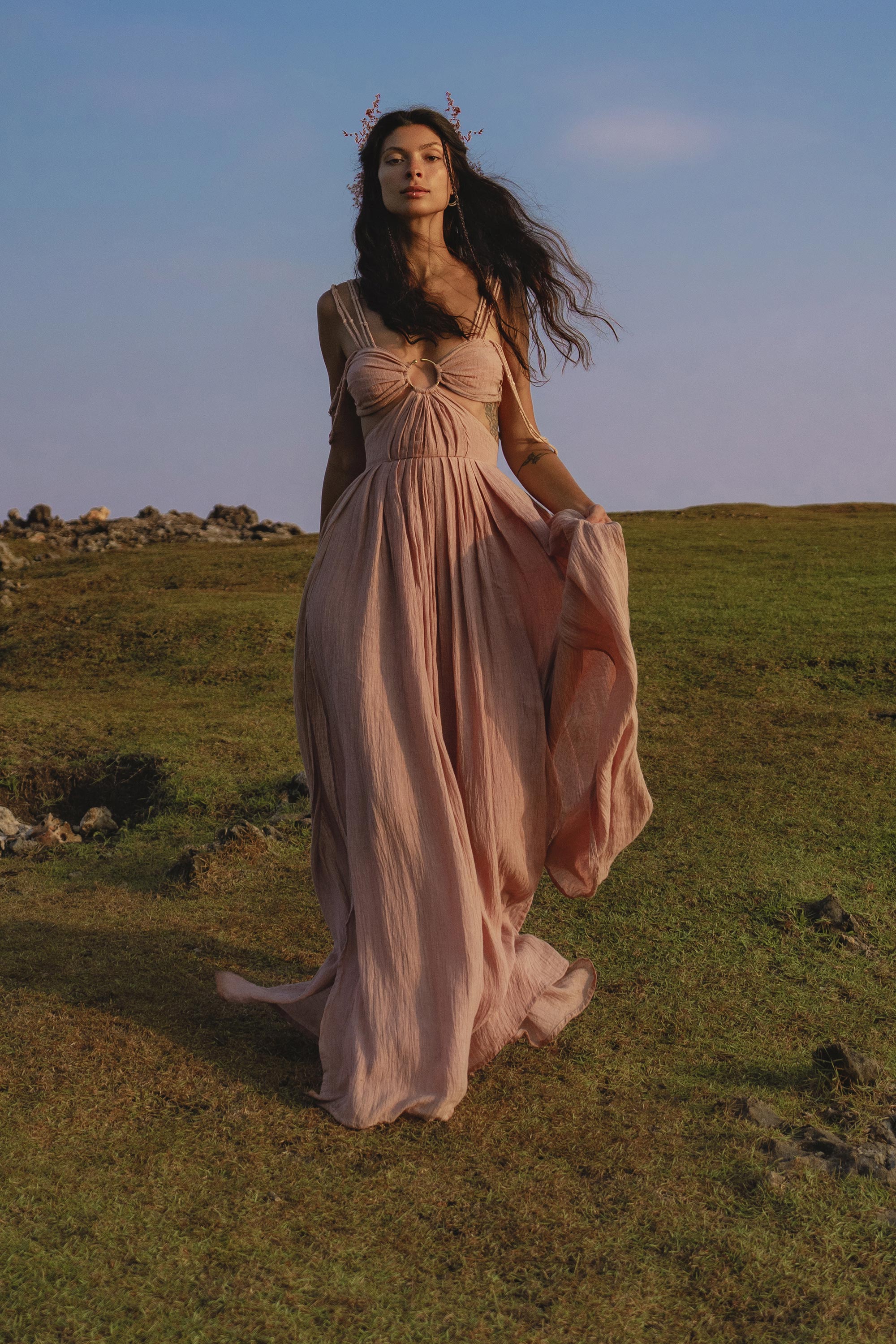 A woman with long dark hair strolls on grass beneath a blue sky, wearing the Ananda Lālita Dress Blush Rose – Crinkle Linen Goddess Dress by AYA Sacred Wear. She holds her flowing dress and wears floral headpieces for an ethereal look.