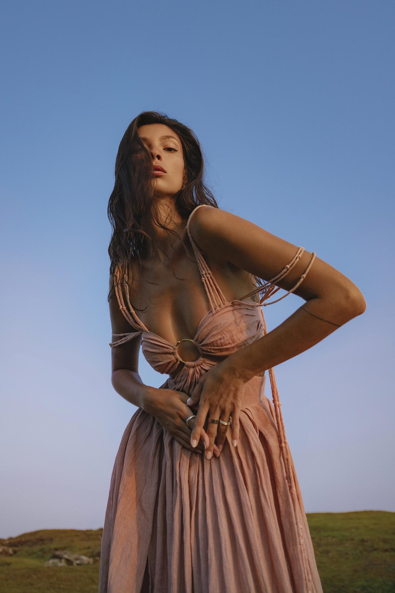 A woman with long dark hair stands confidently on green grass under a blue sky, wearing the Ananda Lālita Dress Blush Rose – Crinkle Linen Goddess Dress by AYA Sacred Wear, styled with handcrafted brass rings and a serene expression.