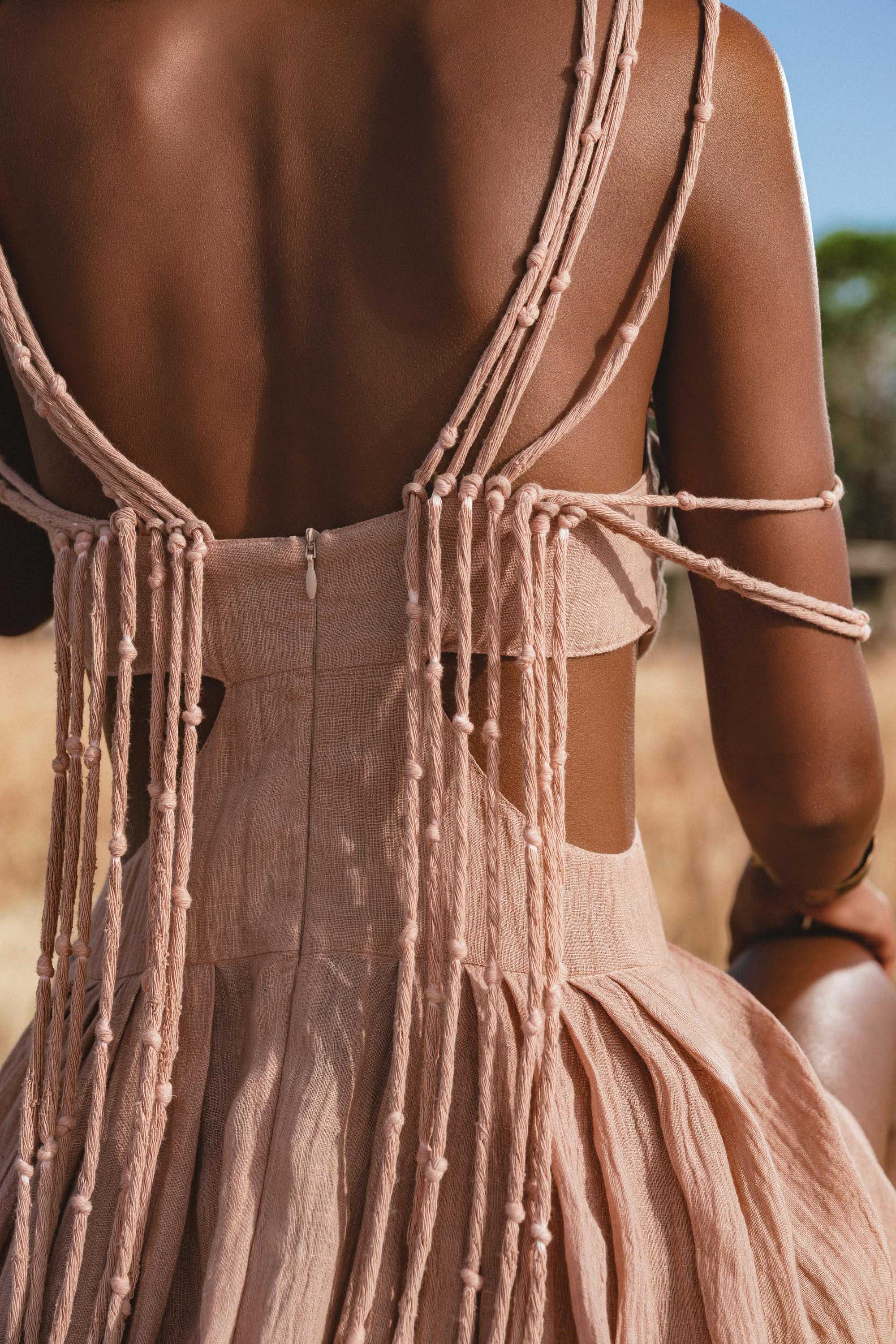 A close-up shows a woman’s back in the Ananda Lālita Dress Blush Rose – Crinkle Linen Goddess Dress by AYA Sacred Wear, featuring knotted draped strings and handcrafted brass rings, glowing in sunlight amid dry grass and greenery.