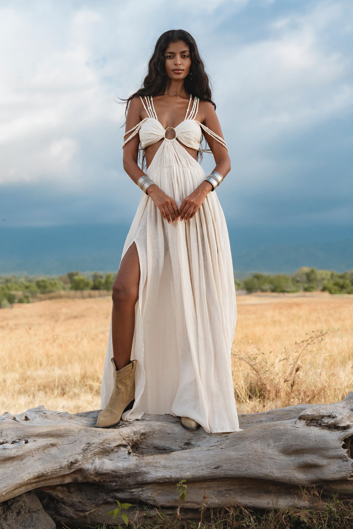 A woman stands outdoors on a log in a golden field, wearing the Ananda Lālita Dress in ivory by AYA Sacred Wear. She pairs the linen organic goddess gown with silver cuffs, brass rings, beige ankle boots, and wears her long dark hair loose. Mountains rise behind.