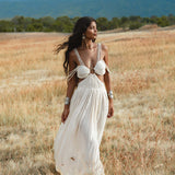 A woman in the Ananda Lālita Dress Ivory Colour – Linen Organic Goddess Gown by AYA Sacred Wear strolls through a sunlit grassy field, her long dark hair flowing and handcrafted brass rings gleaming, with mountains and clouds in the background.