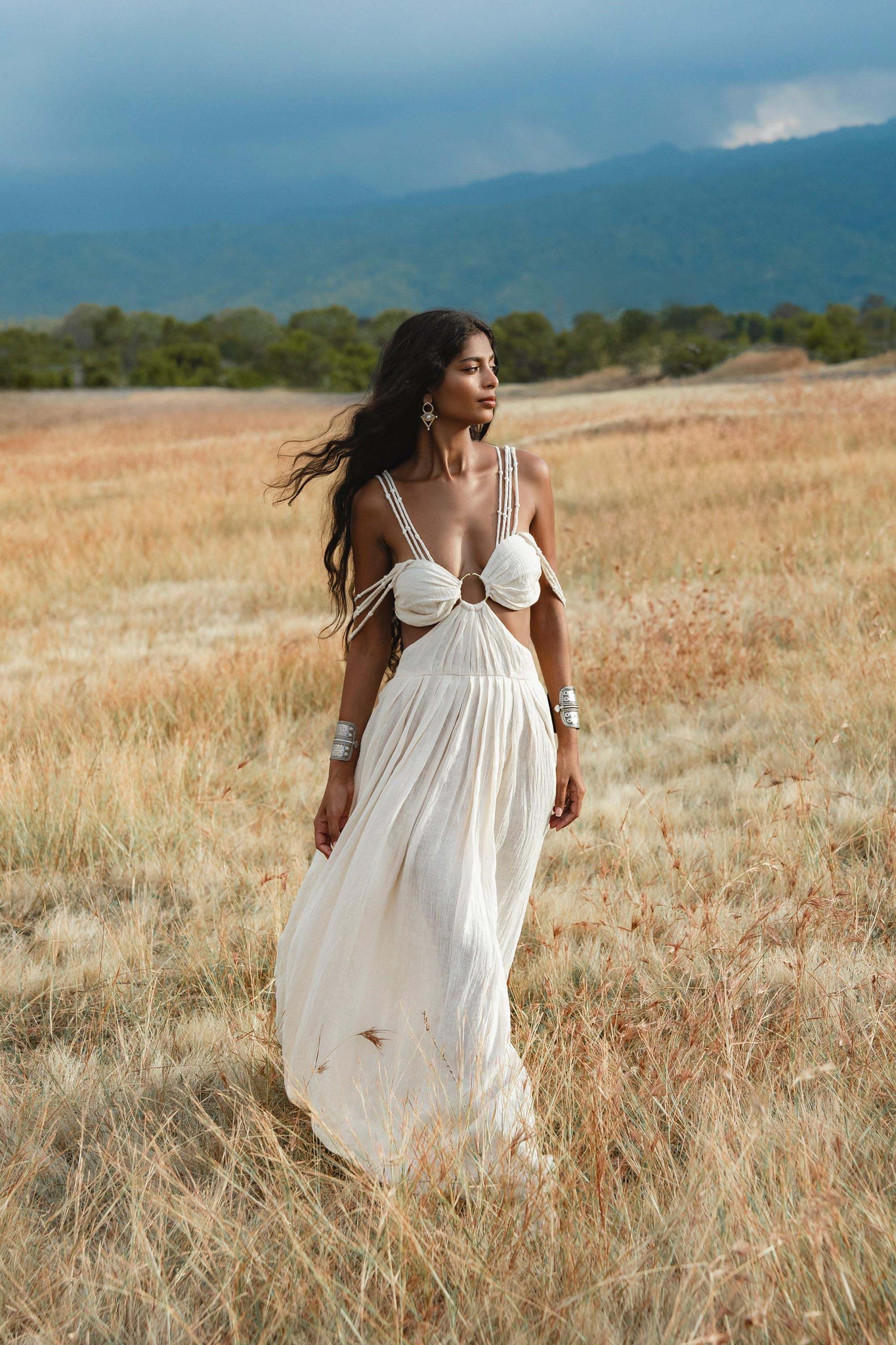 A woman in the Ananda Lālita Dress Ivory Colour – Linen Organic Goddess Gown by AYA Sacred Wear strolls through a sunlit grassy field, her long dark hair flowing and handcrafted brass rings gleaming, with mountains and clouds in the background.