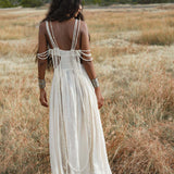 A woman with long wavy dark hair stands in a dry grassy field, facing away. She wears the Ananda Lālita Dress in ivory linen by AYA Sacred Wear, featuring beaded drapes over her arms and back, paired with brass rings and silver bracelets.