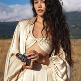 A woman with long, wavy dark hair stands in a sunlit field, wearing the AYA Sacred Wear Apsara Bra in Cream Colour—pure silk, handcrafted in Bali. She holds a vintage camera as mountains and blue sky fill the background.