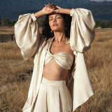 In a dry grassy field with mountains behind her, a woman shields her face from the sun, eyes closed. She wears the AYA Sacred Wear Apsara Bra in Cream Colour—pure silk and transformable, inspired by handcrafted Balinese styles.