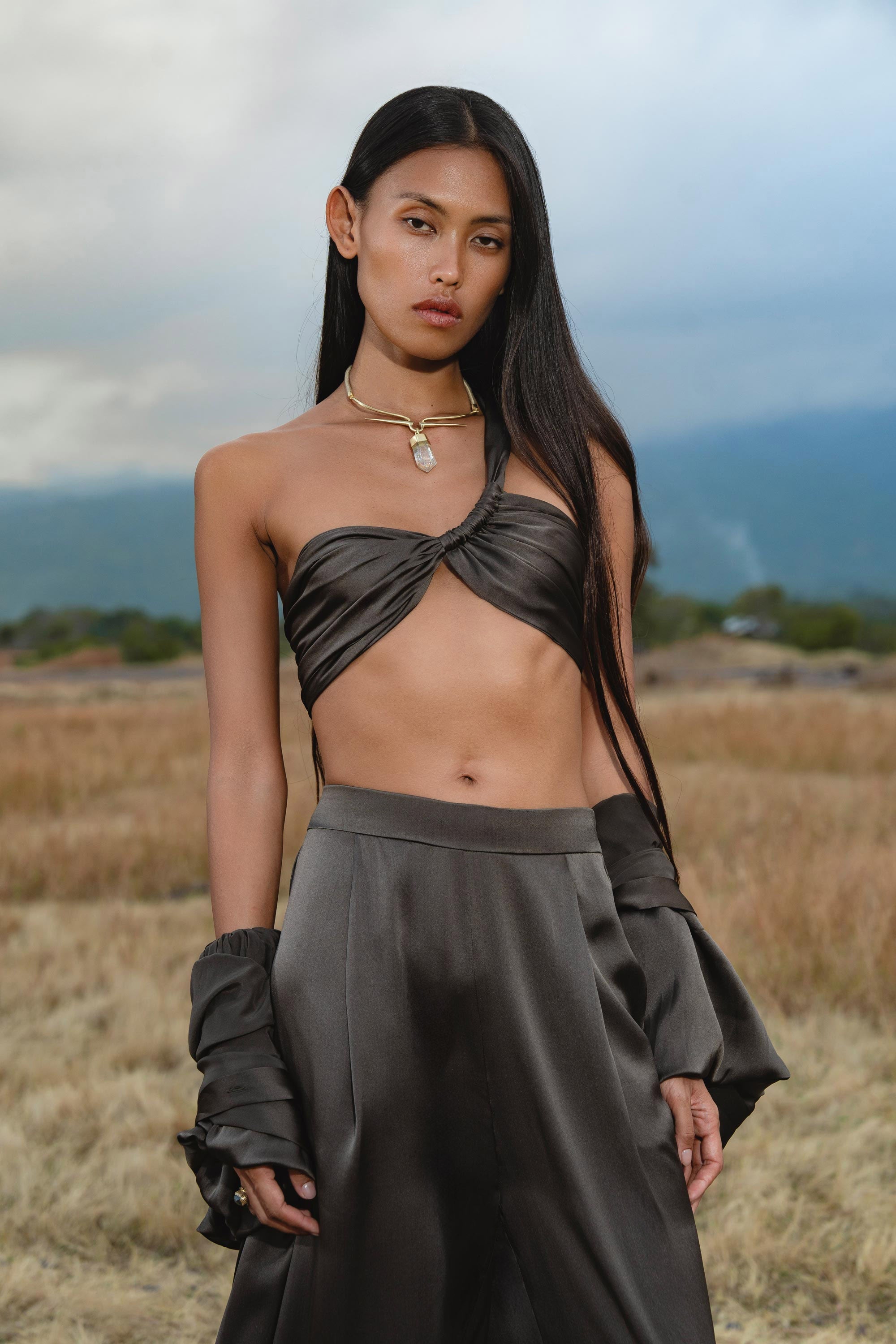 A person with long, straight black hair stands in a grassy field, wearing the Apsara Bra in Asphalt Gray – Pure Silk Transformable by AYA Sacred Wear and flowy pants. They accessorize with a gold necklace and bracelet under cloudy skies with distant mountains.