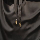 A close-up of the AYA Sacred Wear Apsara Bra in Asphalt Gray – Pure Silk Transformable shows smooth, shiny silk fabric and two black strings tied in a knot, each finished with a handcrafted gold wing charm for elegant detail.