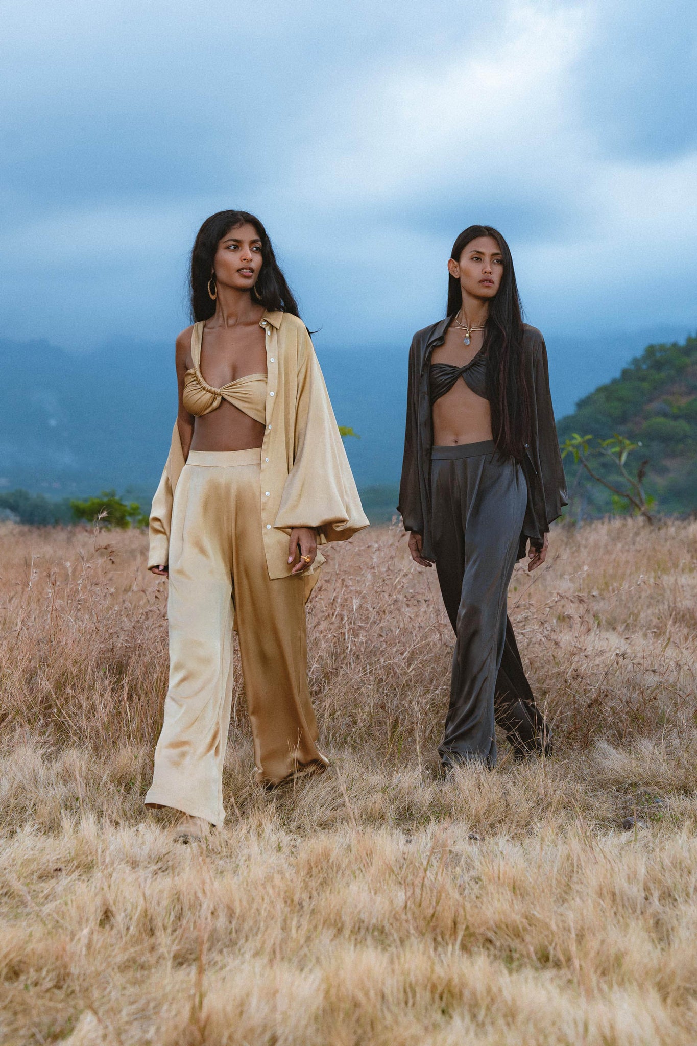 Two women walk side by side in a dry field with mountains behind them. One wears the Apsara Bra in Asphalt Gray by AYA Sacred Wear—pure silk, transformable, and handcrafted in Bali for effortless style and artistry.