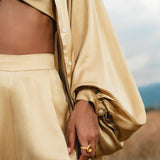 Wearing the AYA Sacred Wear Apsara Bra in Inka Gold—pure silk and transformable—a person outdoors touches their side, accessorized with a chunky gold ring, completing a chic look of bandeau top, high-waisted pants, and voluminous-sleeved shirt.