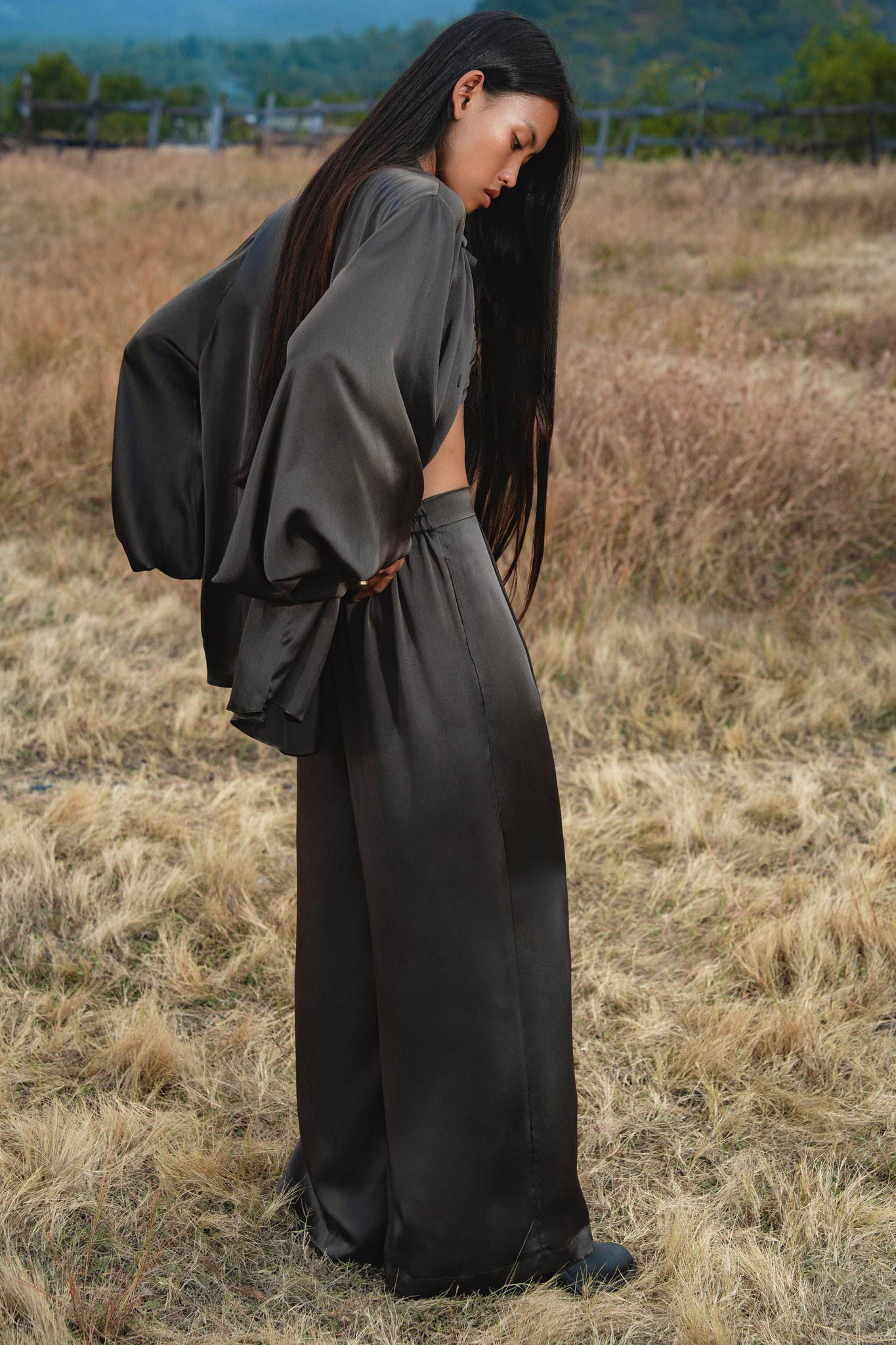A woman with long dark hair stands in a dry grassy field, wearing loose black satin pants and the AYA Sacred Wear Apsara Kimono Blouse in Asphalt Gray—a pure silk kimono handcrafted in Bali. She gazes downward, hills softly blurred behind her.