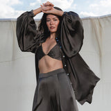 A woman with long dark hair poses outdoors against a fabric backdrop, wearing the AYA Sacred Wear Apsara Kimono Blouse in Asphalt Gray pure silk over a black bralette and wide-leg pants, confidently gazing at the camera.