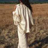 A person with long, wavy dark hair stands in a dry grassy field wearing the Apsara Kimono Blouse Cream – Pure Silk Kimono by AYA Sacred Wear. Green trees and distant mountains rise under a partly cloudy sky in the background.