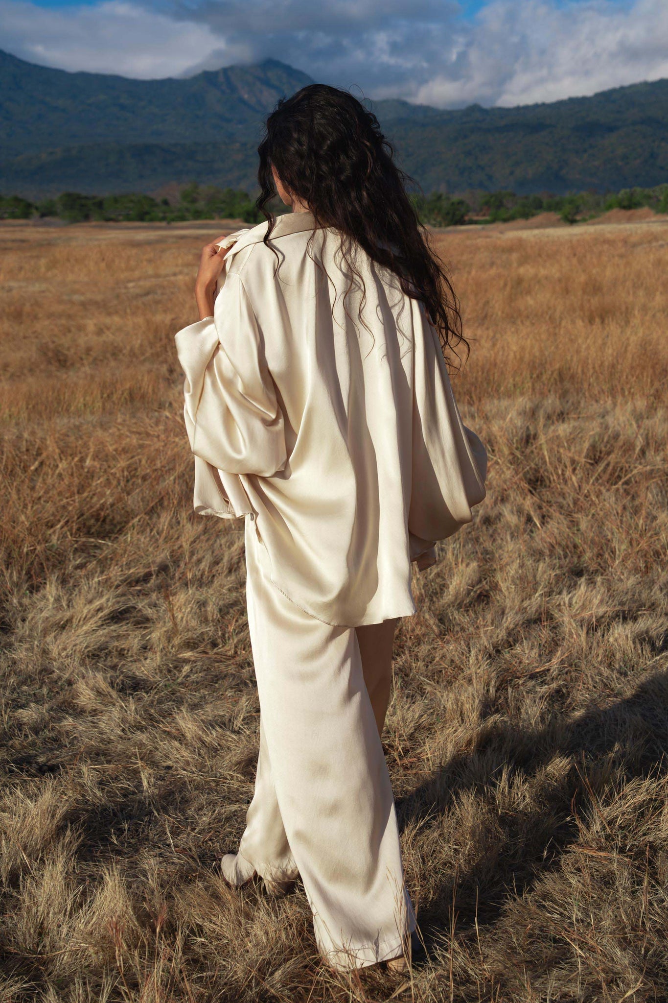 A person with long, wavy dark hair stands in a dry grassy field wearing the Apsara Kimono Blouse Cream – Pure Silk Kimono by AYA Sacred Wear. Green trees and distant mountains rise under a partly cloudy sky in the background.