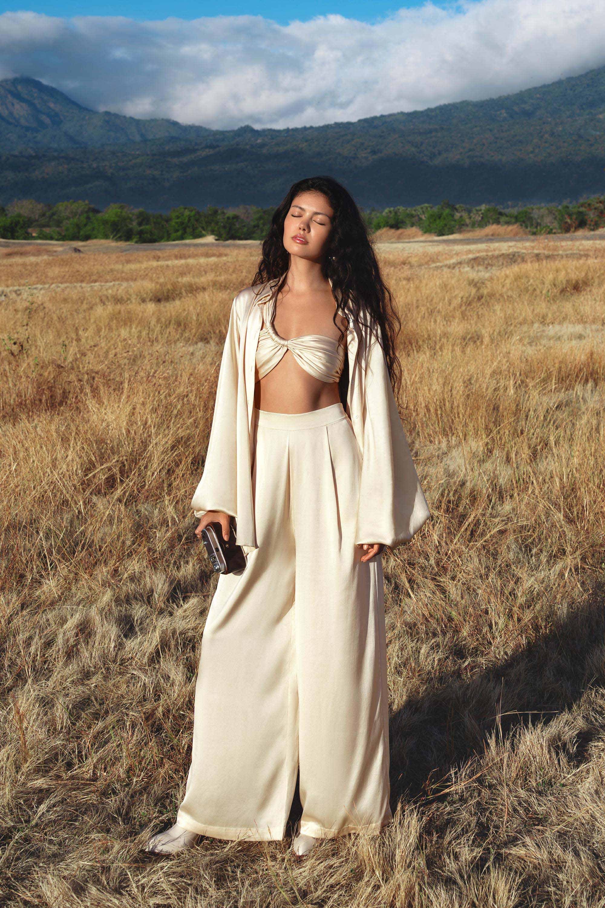 A woman with long wavy hair stands in a dry grassy field, wearing the AYA Sacred Wear Apsara Kimono Blouse Cream – a pure silk kimono handcrafted in Bali. She pairs it with wide-legged pants and a bralette, gazing serenely at distant mountains.