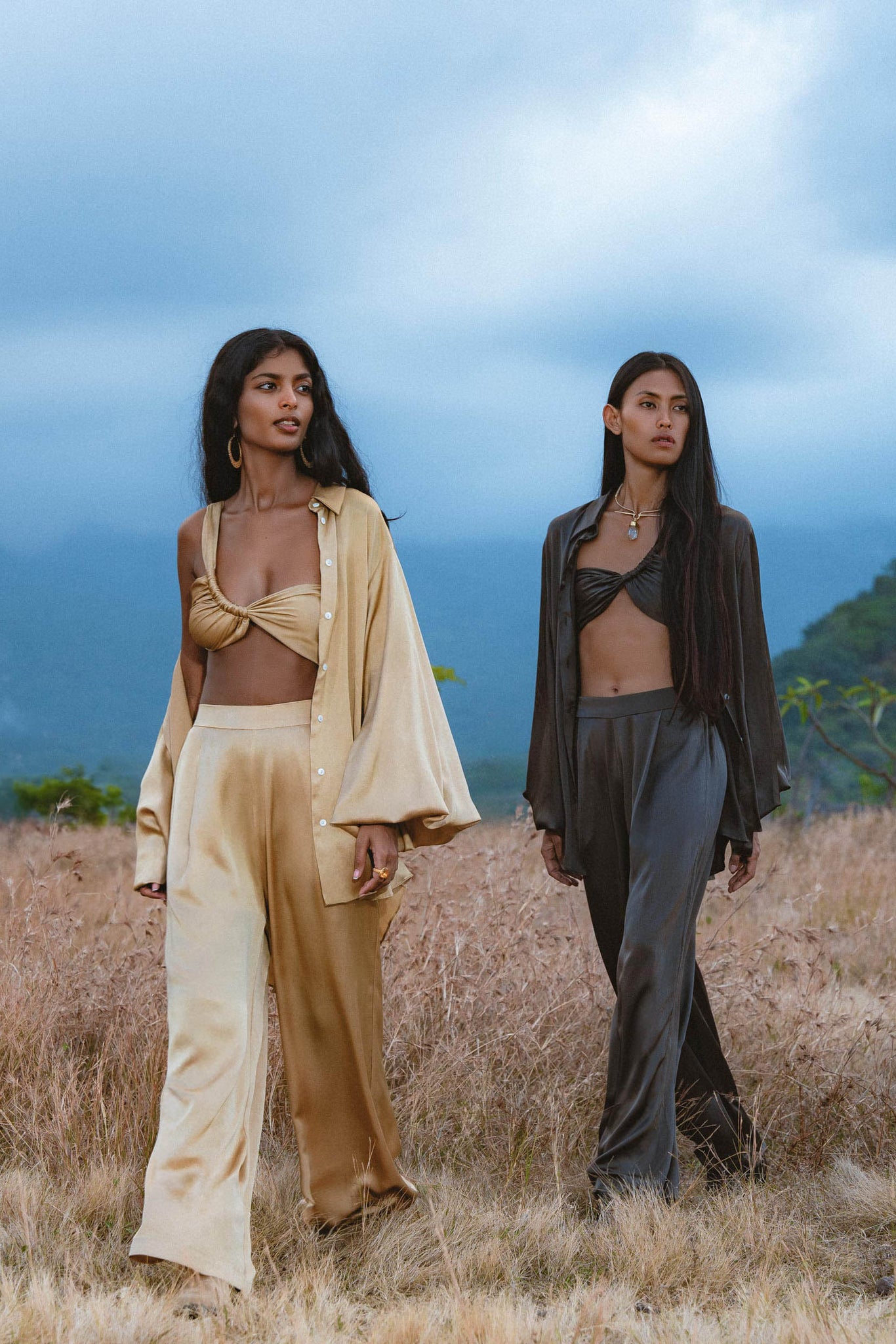 Two women stand in a grassy field with mountains behind. One wears the AYA Sacred Wear Apsara Kimono Blouse Inka Gold, handcrafted from pure silk, paired with wide pants; the other sports a dark brown bralette and matching pants for a calm, stylish look.