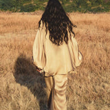 A woman with long, wavy dark hair walks through a golden field, wearing the AYA Sacred Wear Apsara Kimono Blouse Inka Gold— a pure silk top for women— as green hills rise beneath a blue sky, creating a serene natural mood.