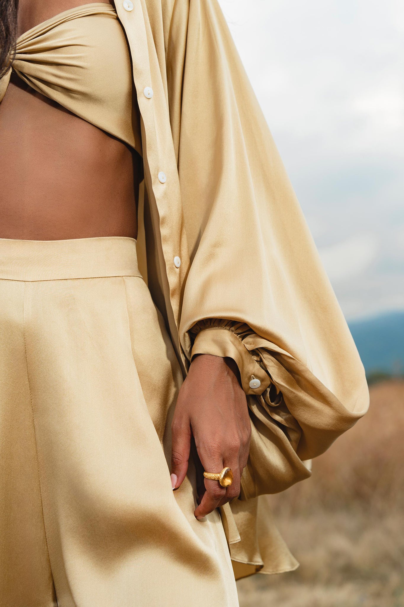 Wearing the AYA Sacred Wear Apsara Kimono Blouse Inka Gold—a pure silk top for women—paired with a bandeau and high-waisted pants, a person stands in a field, gold ring visible, with only their torso and hand shown.