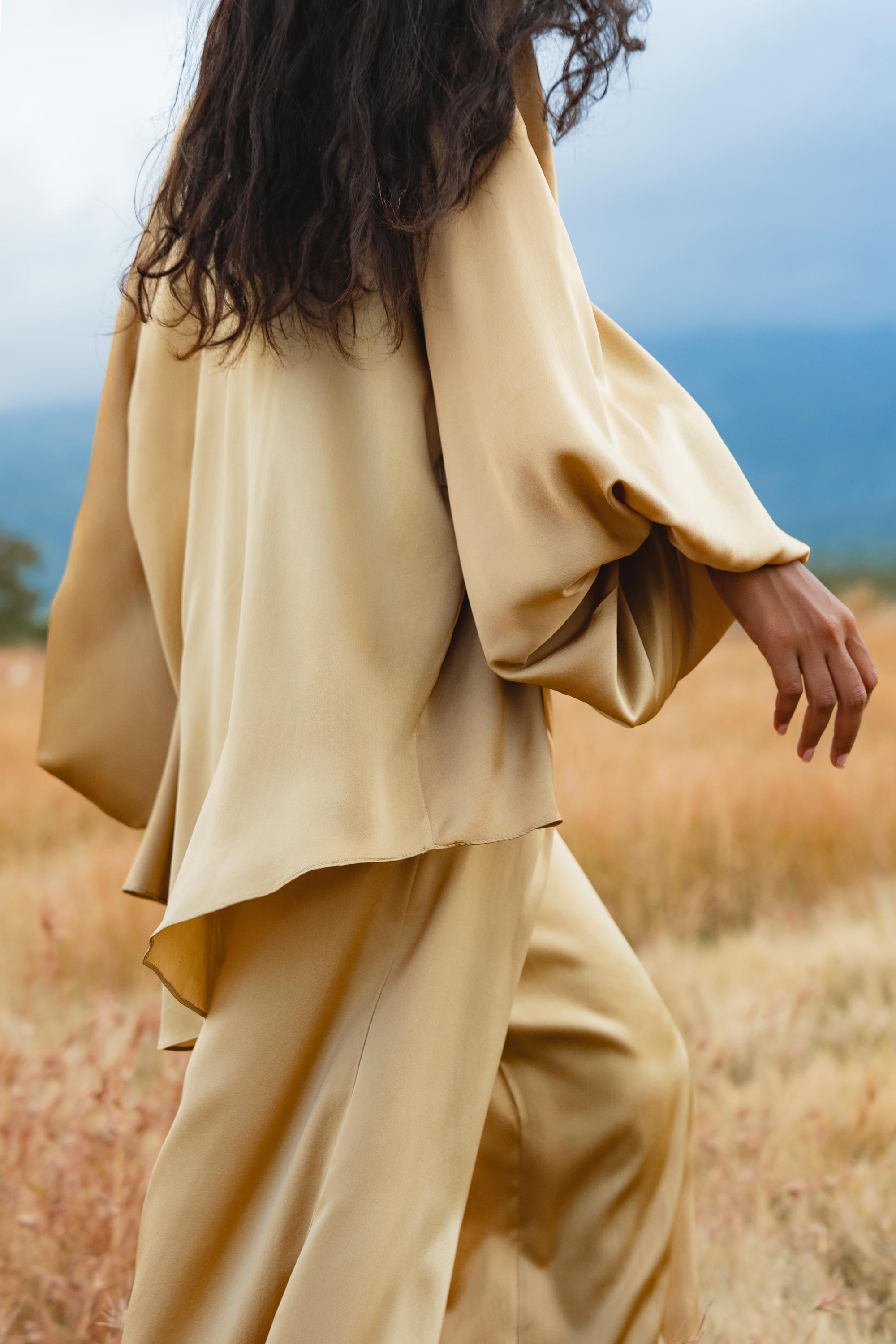 A woman with long, wavy dark hair wears the AYA Sacred Wear Apsara Kimono Blouse Inka Gold – a pure silk top for women – paired with wide pants, walking through tall golden grass under a cloudy sky with distant blue mountains. Face not visible.