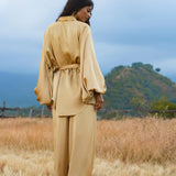 A woman stands in a dry, grassy field wearing the AYA Sacred Wear Apsara Kimono Blouse Inka Gold—a pure silk, loose-fit belted top for women—paired with matching pants, her dark hair down against green hills and a cloudy sky.