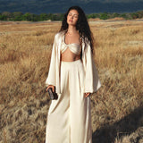 A woman with long, wavy hair stands in a dry grass field, eyes closed. She wears AYA Sacred Wear’s Apsara Pants in cream—pure silk flowy pants—paired with a matching jacket and bandeau, holding a clutch against a mountain backdrop.