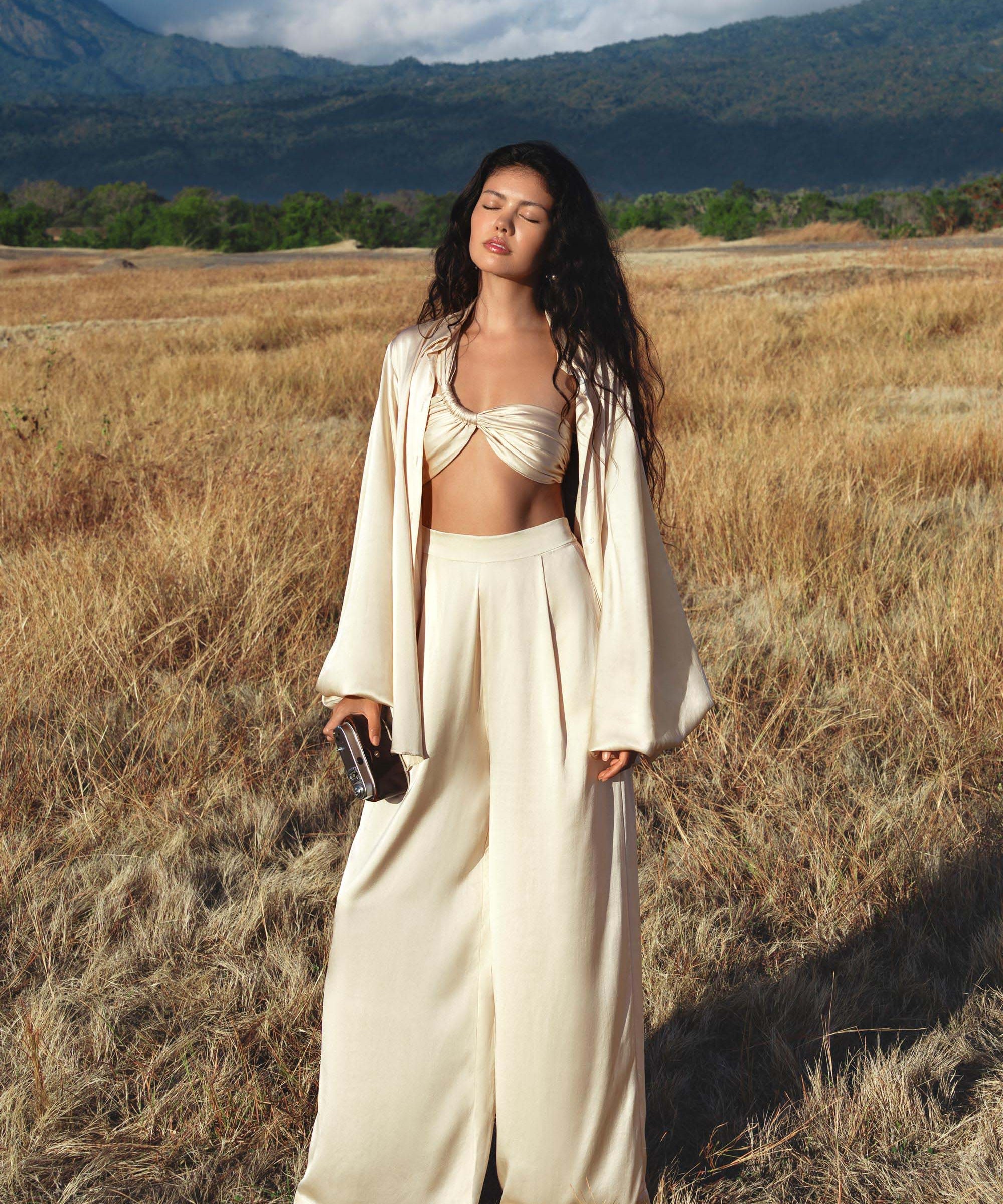 A woman with long, wavy hair stands in a dry grass field, eyes closed. She wears AYA Sacred Wear’s Apsara Pants in cream—pure silk flowy pants—paired with a matching jacket and bandeau, holding a clutch against a mountain backdrop.