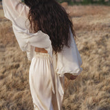 A woman with long wavy hair, dressed in AYA Sacred Wear's Apsara Pants Cream Colour – Pure Silk Flowy Pants, a cream blouse, and a decorated wide-brimmed hat, strolls through a grassy field radiating modern bohemian style.