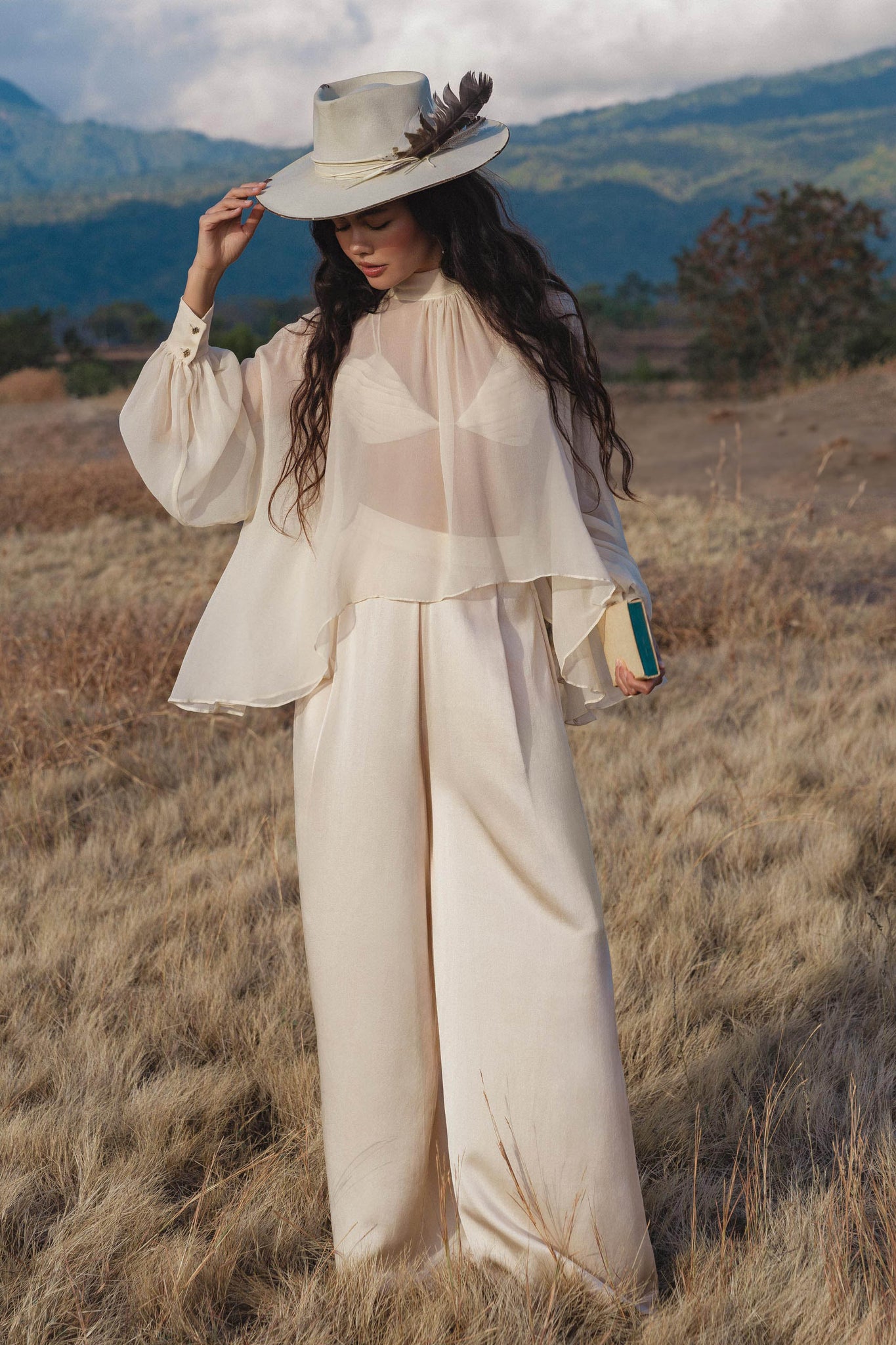 In a dry grassy field with mountain views, a woman in AYA Sacred Wear's Apsara Pants Cream Colour—pure silk flowy pants—pairs them with a sheer blouse and feathered hat. She holds a book, gazing down as her long wavy hair falls over her shoulders.