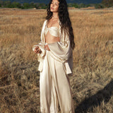 In a sunlit field, a woman in AYA Sacred Wear’s Apsara Pants—cream-coloured, pure silk, and flowy—stands with closed eyes. Her long dark hair cascades as mountains and clouds frame the serene scene.