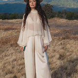 A woman with long wavy hair stands in a grassy field wearing AYA Sacred Wear’s Apsara Pants in cream—pure silk flowy pants—paired with a sheer top and wide-brimmed hat, holding a small book against a mountain and cloud backdrop.