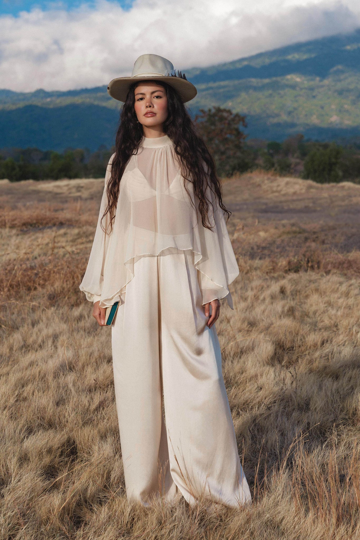 A woman with long wavy hair stands in a grassy field wearing AYA Sacred Wear’s Apsara Pants in cream—pure silk flowy pants—paired with a sheer top and wide-brimmed hat, holding a small book against a mountain and cloud backdrop.