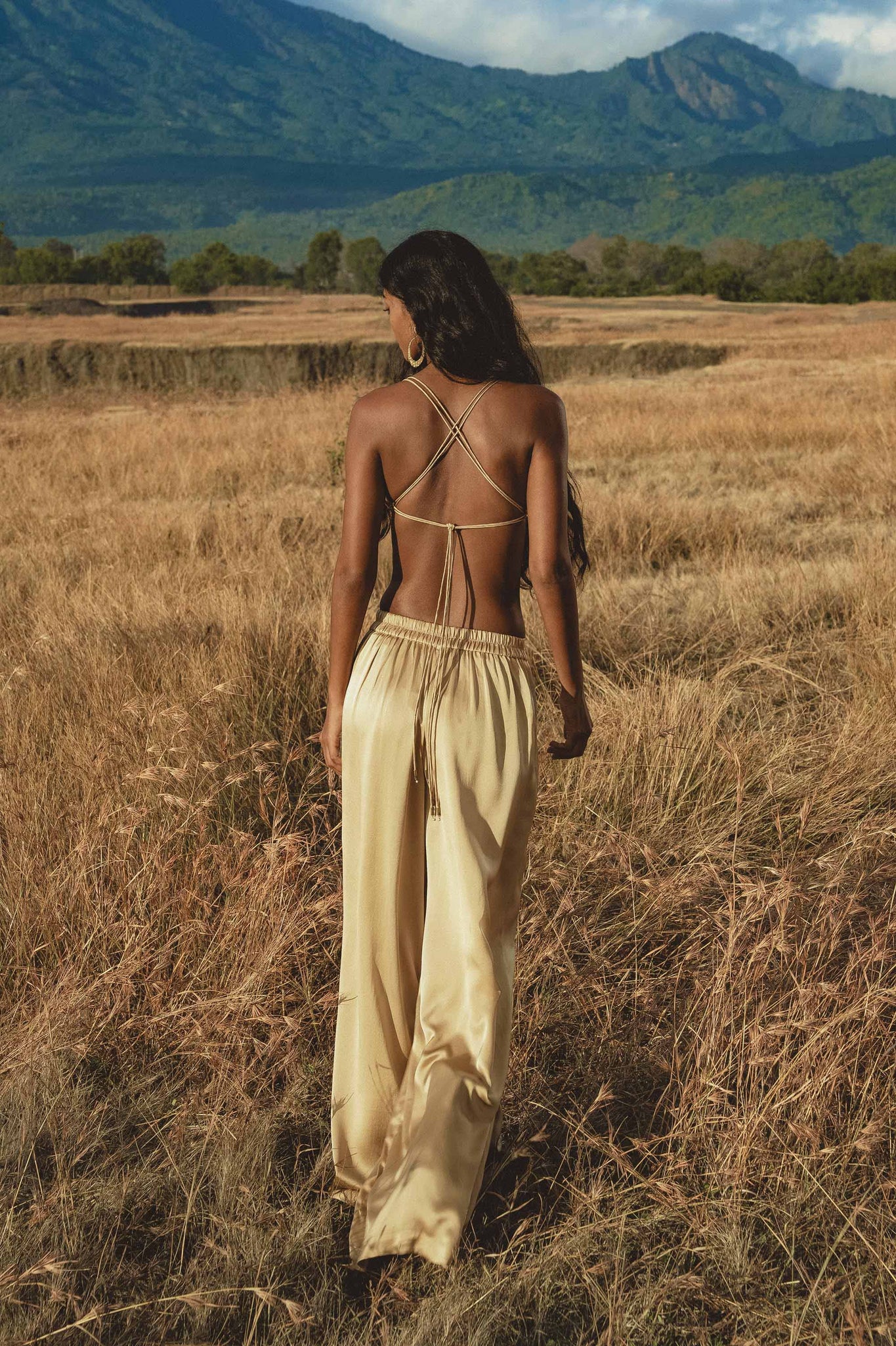 A woman with long dark hair, wearing a beige backless strappy top and AYA Sacred Wear’s Apsara Pants Inka Gold – Pure Silk Flowy Pants, walks through tall golden grass in a field with mountains and trees in the background, facing away from the camera.