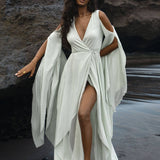 A woman in an AYA Sacred Wear Sky Blue Makosh Goddess Dress, a boho cotton wrap with open shoulders and flowing sleeves, stands barefoot on a dark sandy beach. Her eyes are closed as she leans against a rugged rock, while waves form a harmonious background.