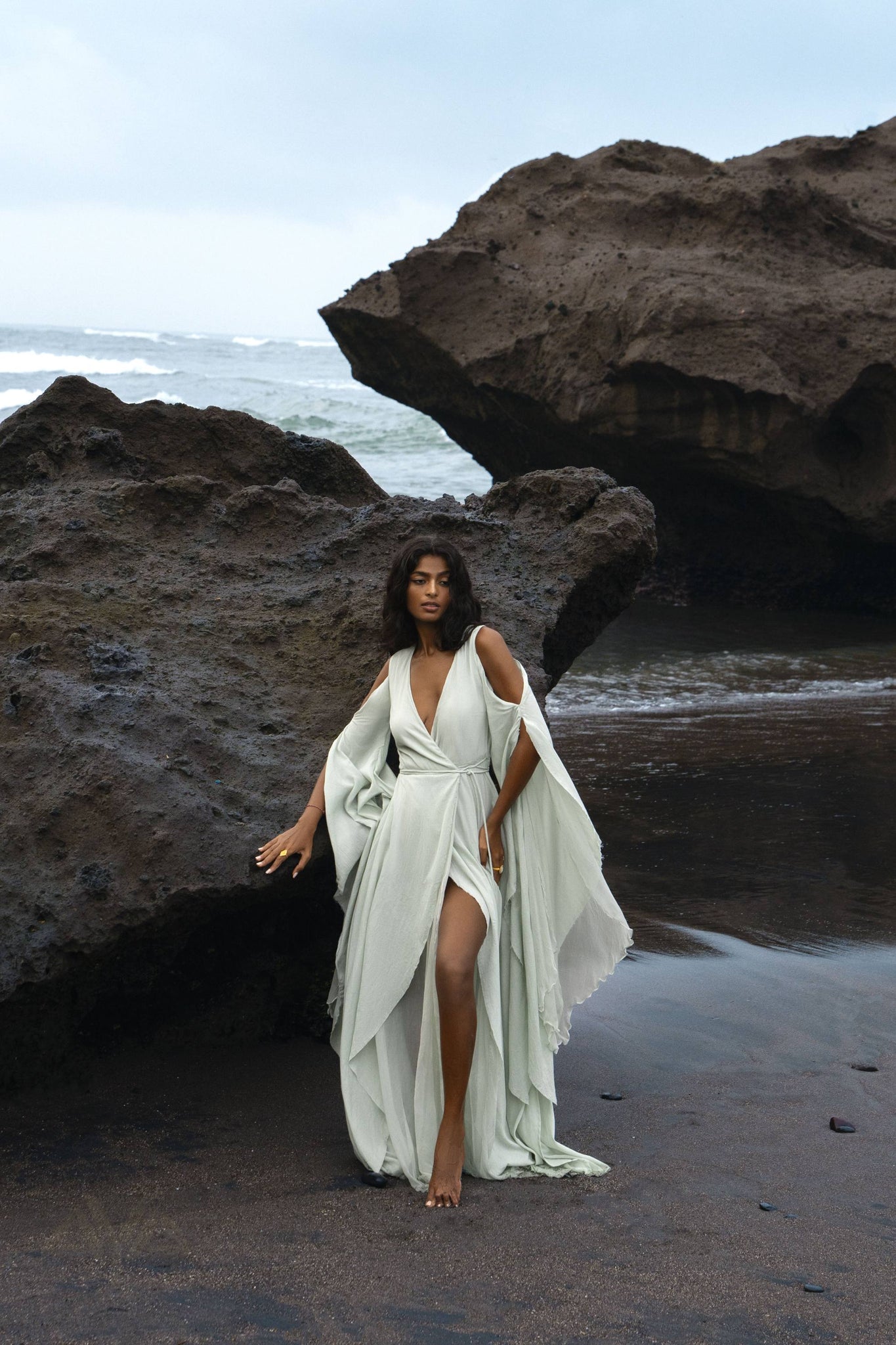 A woman stands barefoot on a rocky beach, wrapped in an AYA Sacred Wear Aquamarine Sundari Goddess Silk Gown with deep V-neck and slit, leaning against a large rock with the ocean and cloudy sky as her backdrop.