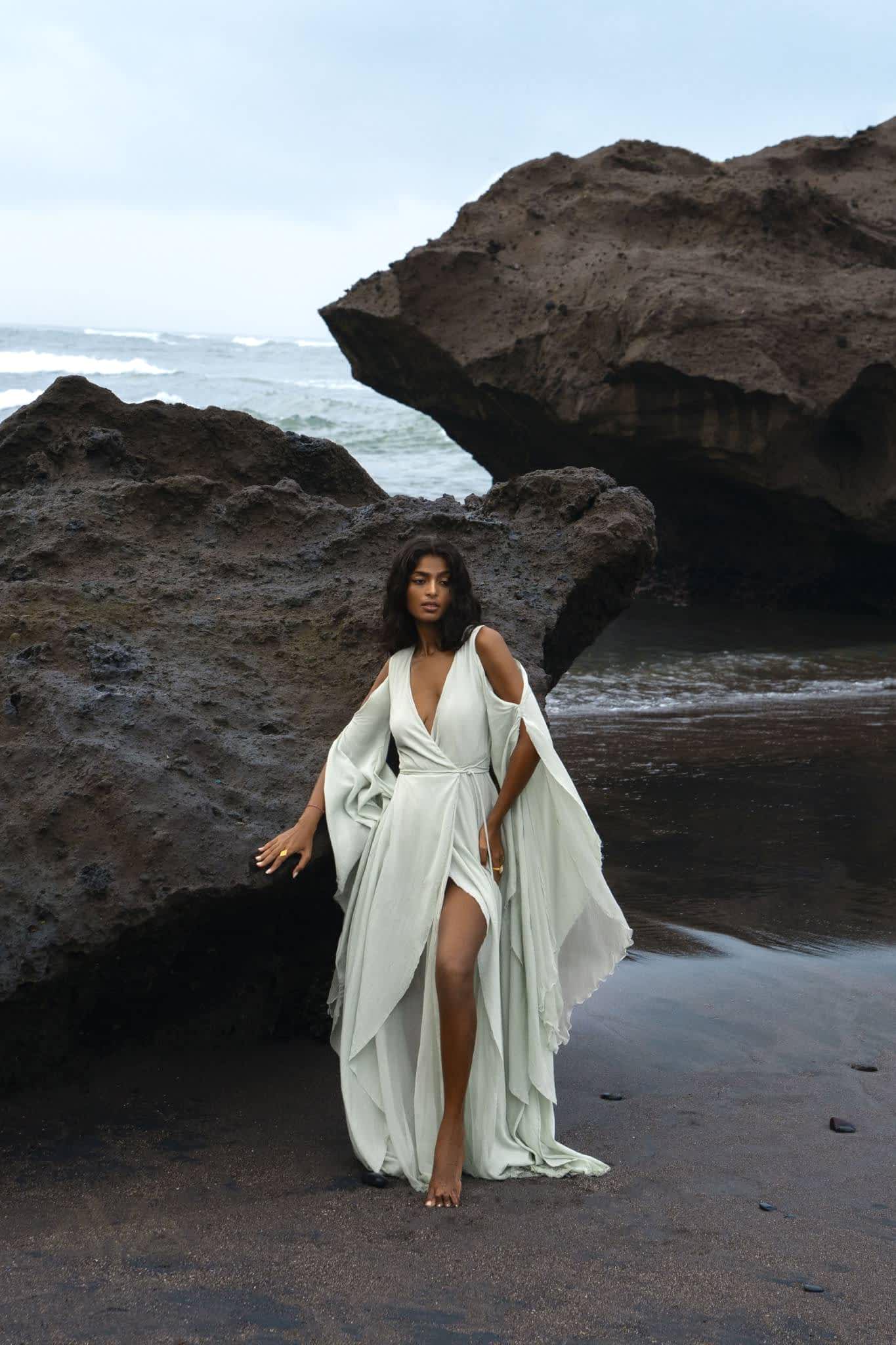 On a rocky beach, a woman in an AYA Sacred Wear Aquamarine Sundari Goddess Silk Gown, featuring ahimsa silk, a deep V-neck, and slit stands barefoot against dark rocks with waves in the background under an overcast sky.