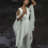 A woman wearing the Aquamarine Sundari Goddess Silk Gown by AYA Sacred Wear gracefully poses on rocky terrain, eyes gently closed. Raising a hand to her head, she exudes serenity as a shimmering pool of water at her feet enhances the contemplative mood.