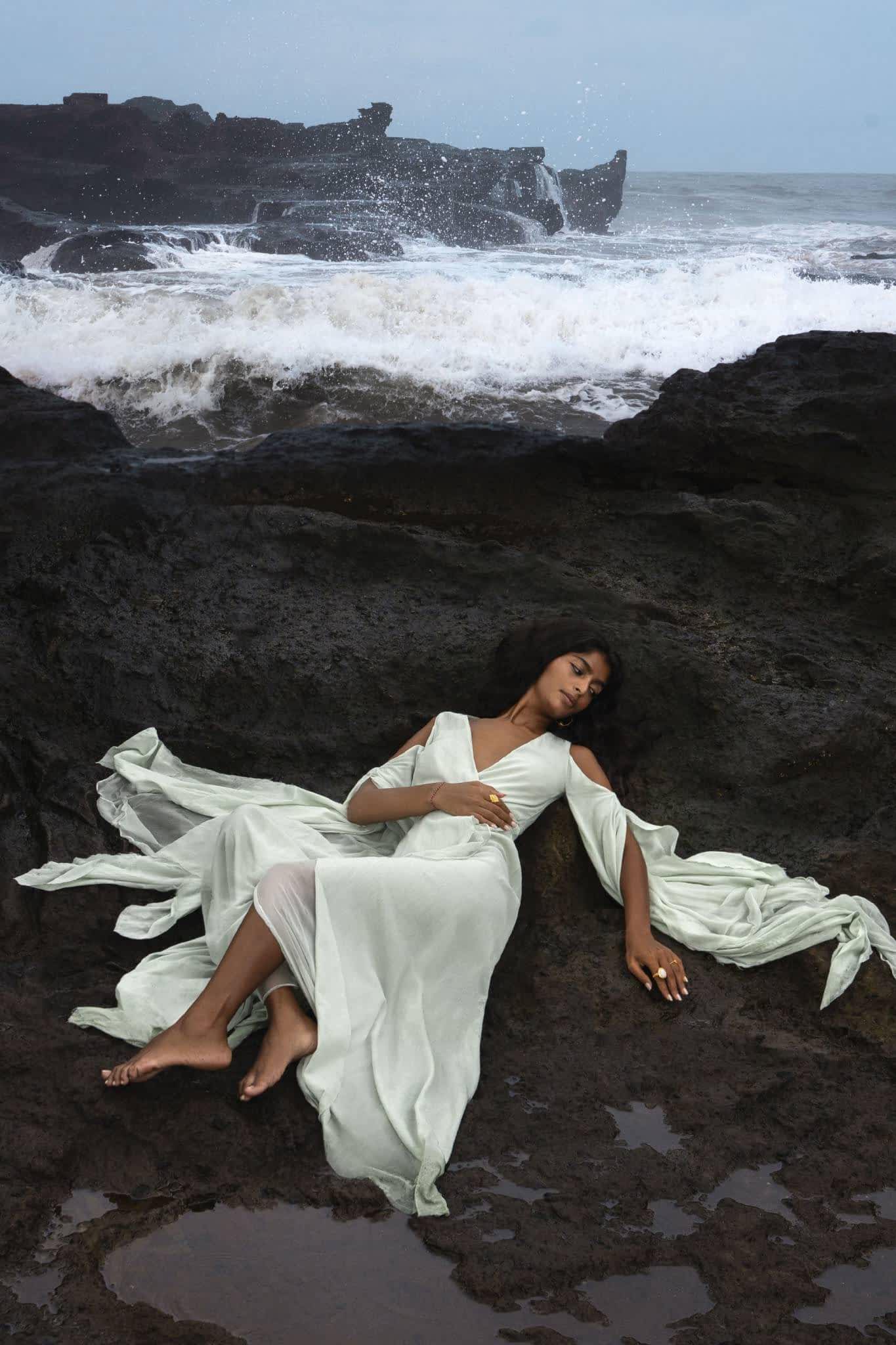 A woman in an AYA Sacred Wear Aquamarine Sundari Goddess Silk Gown lies gracefully on dark, jagged rocks by the sea as waves crash in the background under a cloudy sky, creating a dramatic coastal scene.