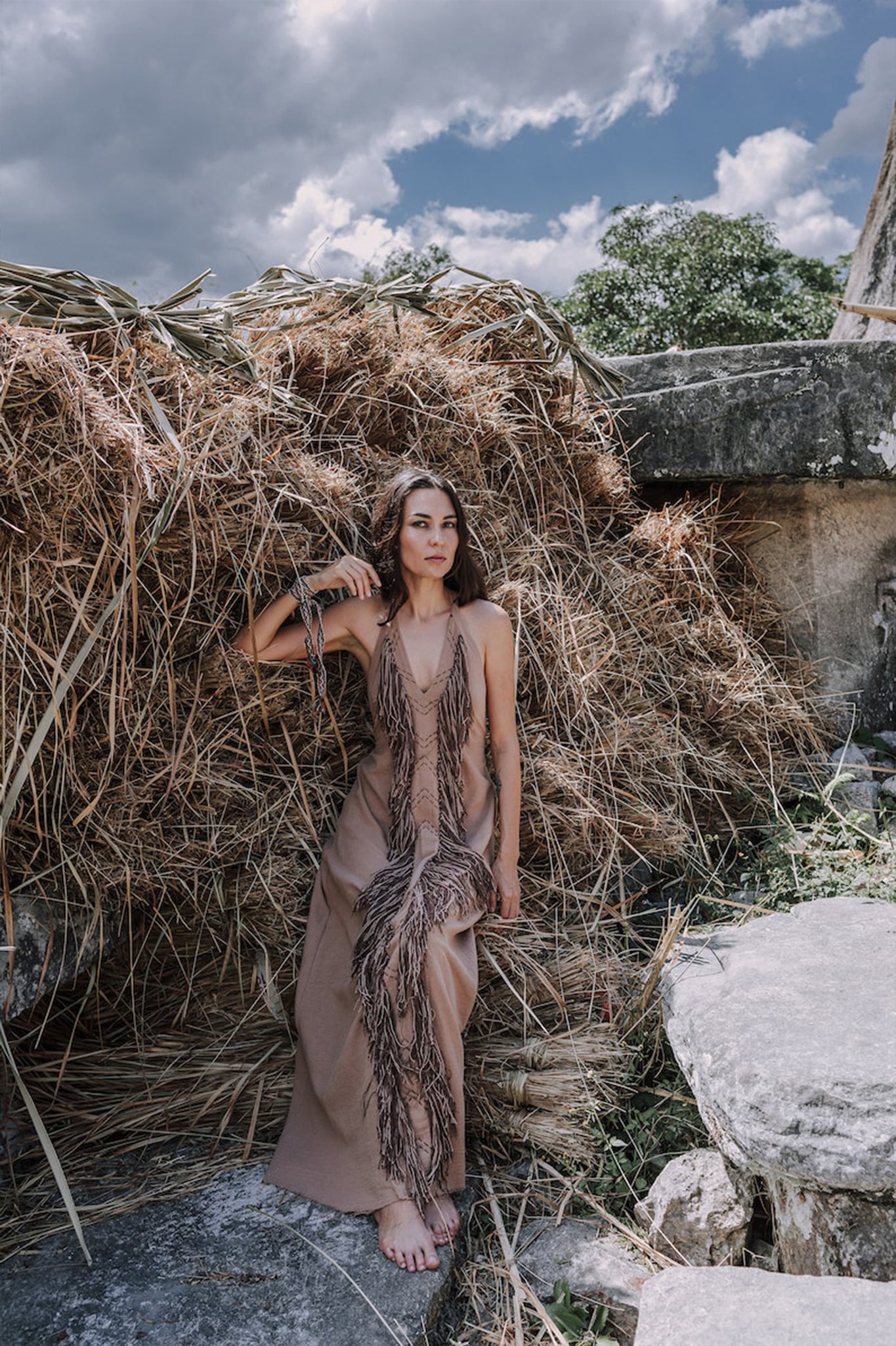 A woman wearing a Beige Organic Cotton Bohemian Tribal Maxi Dress with Hand Loomed Tassels from AYA Sacred Wear sits barefoot on stone steps in front of a large stack of hay. The sky above is partly cloudy, and trees can be seen in the background.